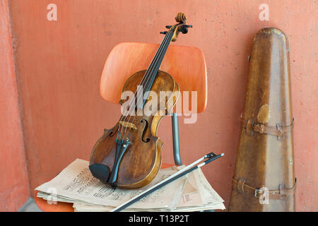 Violin, bow and sheet music on wooden chair with violin case in the background Stock Photo