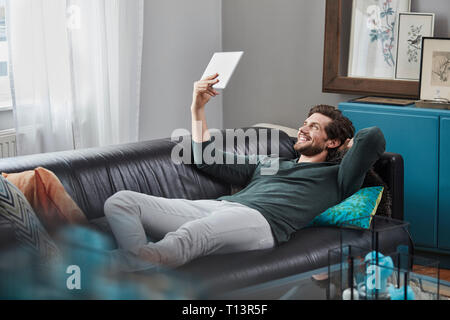 Smiling man with tablet lying on couch at home Stock Photo