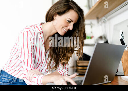 Smiling woman using laptop in kitchen at home Stock Photo