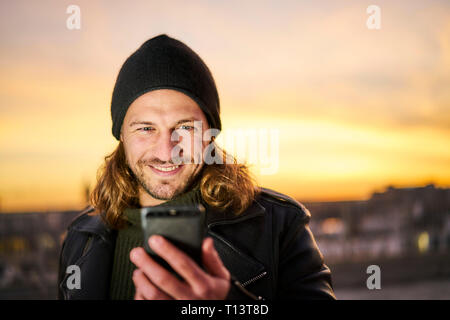 Portrait of bearded young man wearing wooly hat looking at mobile phone by sunset Stock Photo