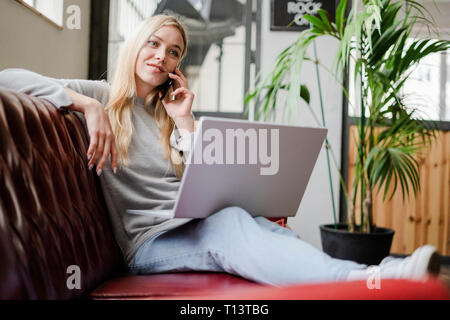 Smiling young woman sitting on couch with laptop talking on cell phone Stock Photo