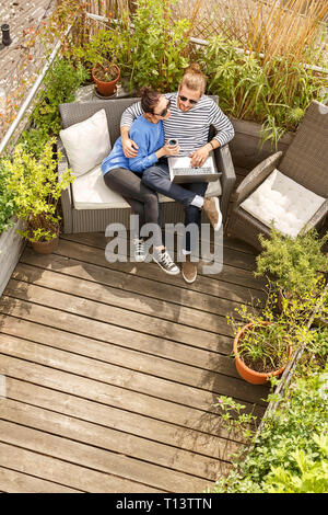Young couple relaxing on their balcony, sitting on couch, using laptop Stock Photo