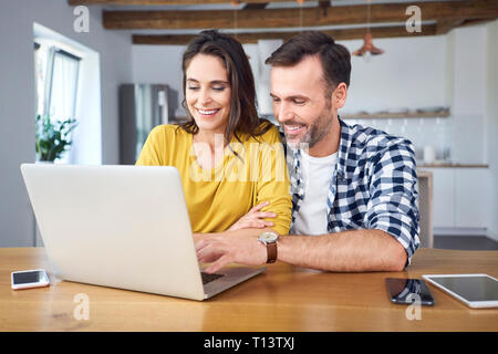 Couple sitting at dining table, using laptop, smiling Stock Photo