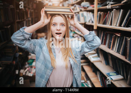 Funny picture of girl standing near bookshelf. She is holding two books on her head with her hands and looking straight forward. She looks amazed Stock Photo