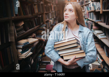 A small girl standing in a big old book store and holding many books in her hands. She is searching for another book to take with her. Young beauty lo Stock Photo