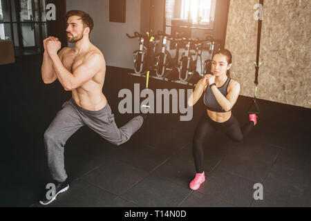 Attractive and slim woman and man are doing lunges on one leg and keeping hands together in front of the body. They are doing this exercise slow but Stock Photo