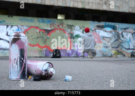 Several used spray cans with pink and white paint lie on the asphalt against the standing guy in front of a painted wall in colored graffiti drawings Stock Photo