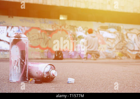 Several used spray cans with pink and white paint lie on the asphalt against the standing guy in front of a painted wall in colored graffiti drawings Stock Photo