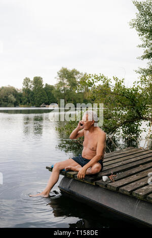 Senior man sitting on jetty at a lake talking on cell phone Stock Photo