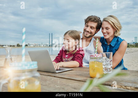 Germany, Duesseldorf, happy family with daughter using laptop on wooden table at Rhine riverbank Stock Photo