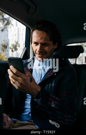 Smiling man sitting on back seat of a car using cell phone Stock Photo