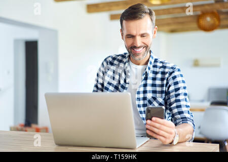 Cheerful man using phone while working on laptop at home Stock Photo