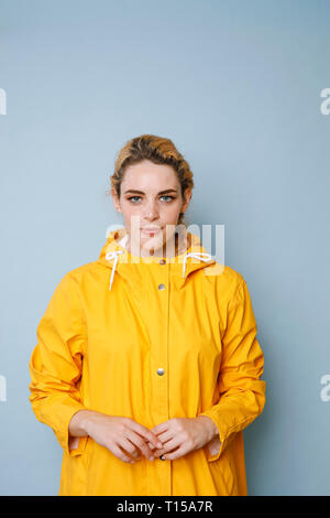 Portrait of young woman wearing yellow rain coat in front of blue background Stock Photo