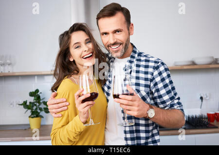 Happy couple standing in kitchen, with arms around, drinking red wine Stock Photo