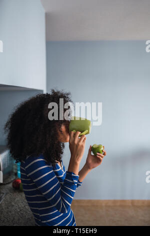 Young woman drinking coffee and eating an apple in her kitchen Stock Photo