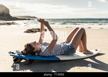 Young woman on the beach, relaxing on surfboard, using smartphone Stock Photo