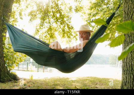 Senior man wearing straw hat relaxing in hammock at lakeshore Stock Photo