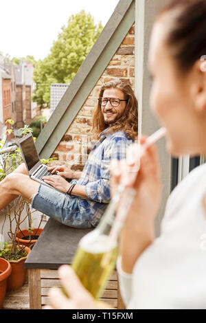 Young couple sitting on their balcony in summer, man using laptop Stock Photo
