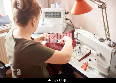 Woman sitting on her workplace in atelier. Female hands using scissors for cutting off fabric piece in sewing process. Overlock, sewing-machine, table Stock Photo