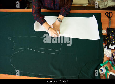 Tailor worktable in the studio. Close-up of female hands making chalk markings of paper patterns on green fabric copy space surface before cutting det Stock Photo