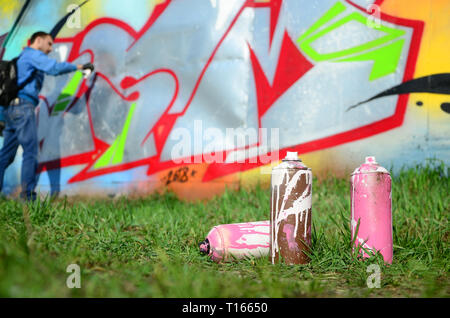 A few used paint cans against the background of the space with the wall on which the young guy draws a large graffiti drawing. Modern art of drawing w Stock Photo