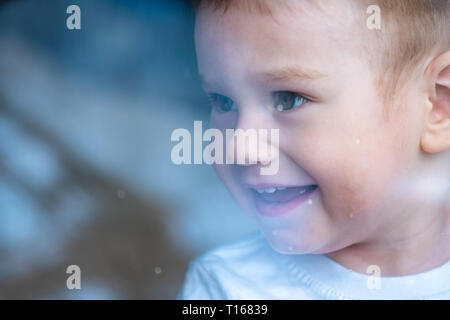 Cute baby boy looking in the window glass with reflection. The concept of loneliness of children and waiting for kindness. Orphanage and orphans Stock Photo