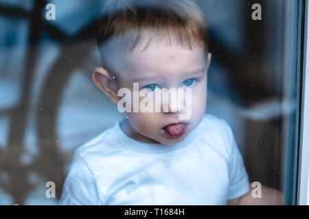 Cute baby boy looking in the window glass with reflection. The concept of loneliness of children and waiting for kindness. Orphanage and orphans Stock Photo