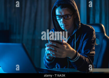 Male hacker in the hood holding the phone in his hands trying to steal access databases with passwords. The concept of cyber security Stock Photo