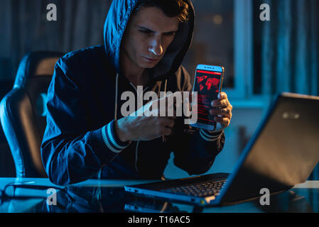 Hacker in the hood holding the phone in his hands trying to steal access databases with passwords. The concept of cyber security Stock Photo