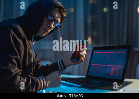 Male hacker in the hood holding the phone in his hands trying to steal access databases with passwords. The concept of cyber security Stock Photo