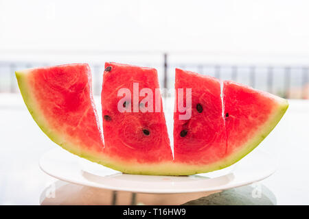 Summer day with vivid vibrant colorful red watermelon slice cut outside on white plate and glass table with seeded black seeds bokeh background of ter Stock Photo