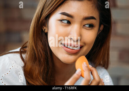 Closeup young Asian positive woman applying powder on shiny face on blurred background Stock Photo