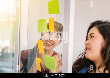 Asian happy businesswomen discussing with sticky notes on window at office Stock Photo