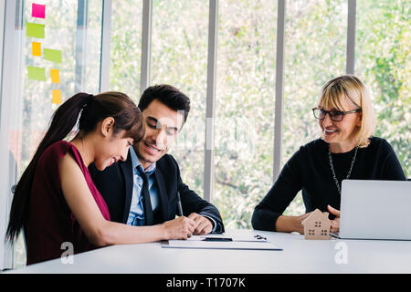 Side view of married couple signing contract and buying house near happy adult agent with laptop at office Stock Photo