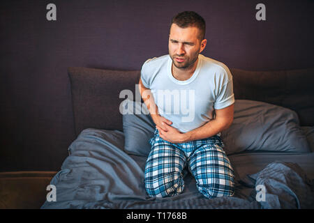 Young man stands on knees on bed and suffers from pain in appendix area. He holds hands there. Guy shrinks. He wear pajamas Stock Photo
