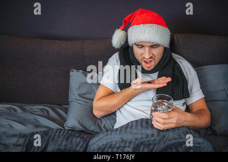 Picture of sick young man going to swallow pills he has on hand. Guy looks at them and shrinks. He has glass of water in another hand. Young man has Stock Photo