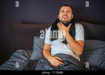 Young man is suffering from strong throat pain. He holds hand close to scarf and shrinks. Guy shrinks and keeps eyes closed. Pain is terrible. Stock Photo