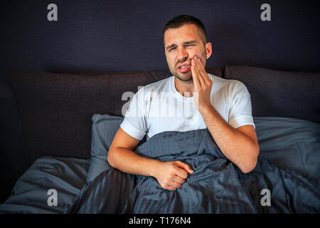 Young man suffering from toothache. He holds hand on cheek and keeps eyes closed. Pain is terrible. He sits on bed. Guy is covered with blanket. Stock Photo