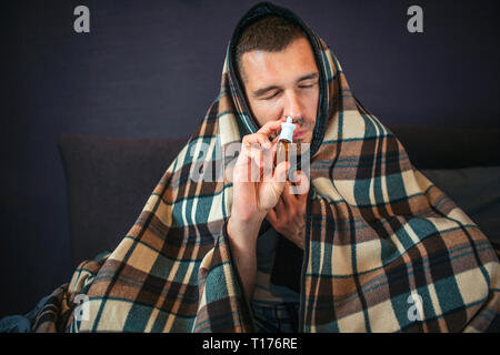 Picture of young man covered all of him with blanket. He uses nose spray. Guy shrinks and keeps eyes closed. Young mean does some tratment for being Stock Photo