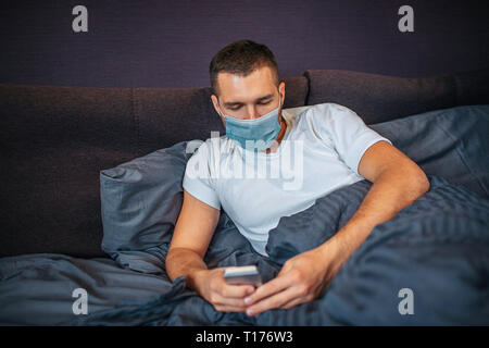 Sick young man is lying on bed and wearing mask. He holds phone in hands and look at it. Guy is calm and concentrated. Young man in in bedroom. Stock Photo