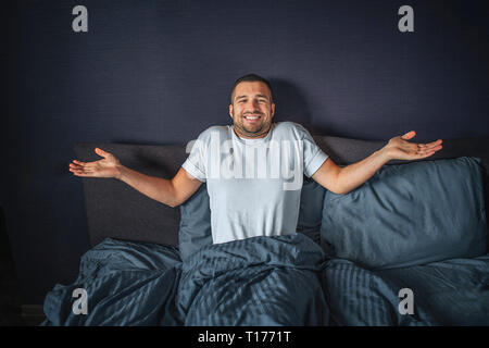 Nice and positive young man sitting on bed. He is covered with dark-blue blanket. He keeps hands aside of body. Guy smiles. He is happy. Stock Photo