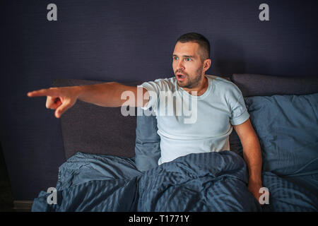 Amazed and wondered young man sitting on bed in bedroom and looks to left. He points with finger. Guy is ecxited. Stock Photo