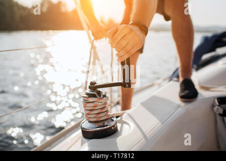 Close up of sailor winds aroung rope by using handle for coiling. He works with both hands. Young man stands on yacht. He is strong. Stock Photo
