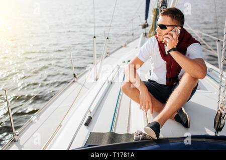 Young man sits on yacht board and look to left. He talks on phone. Guy sits with legs crossed. He wears sunglasses. Young man is busy. Stock Photo