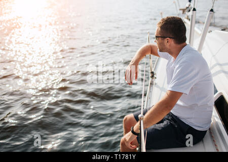 Young man sits at adge of yacht board and looks forward on sun. He leans on railing. Guy is calm and peceful. He is chilling. Water is calm. Stock Photo