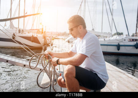 Strong and well-built sailor sits in squad position and holds ropes. He looks at them. Guy is tying knot. Stock Photo