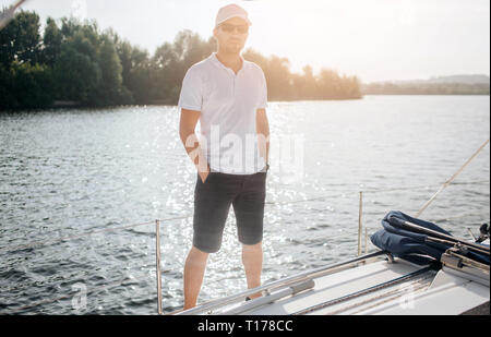 Serious and well-built man stand and pose. He look on camera. Young man wears sunglasses, white shirt and cap. Also guy has dark shorts. Stock Photo
