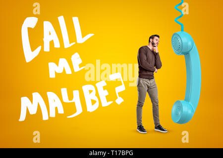 Young man in casual clothes standing in half-turn, one hand on chin, near big cyan phone receiver on yellow background with title Call Me Maybe  Stock Photo