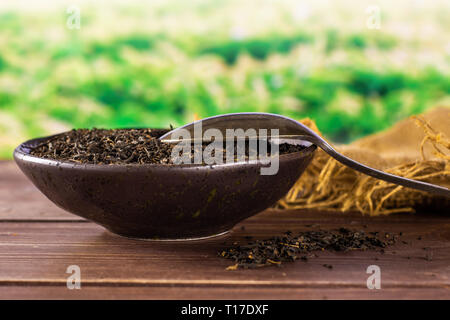 Lot of pieces of dry black tea earl grey in a grey ceramic bowl on jute cloth with silver spoon green tea field behind Stock Photo