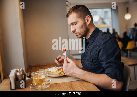 A picture of man sitting at the table and eating. He has a piece of tomato on fork. Guy is looking at it with serious sight Stock Photo